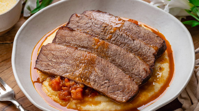 Delicioso brisket assado fatiado sobre polenta cremosa,  acompanhado de molho saboroso, perfeita para uma refeição aconchegante e nutritiva.