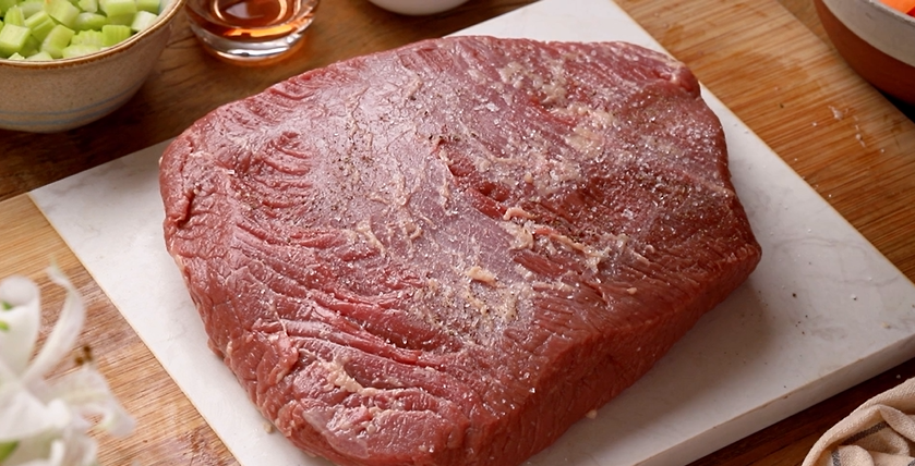 Brisket bovino fresco temperado sobre uma tábua de corte, rodeada por ingredientes como vegetais e molho, ideal para preparo de pratos saborosos.